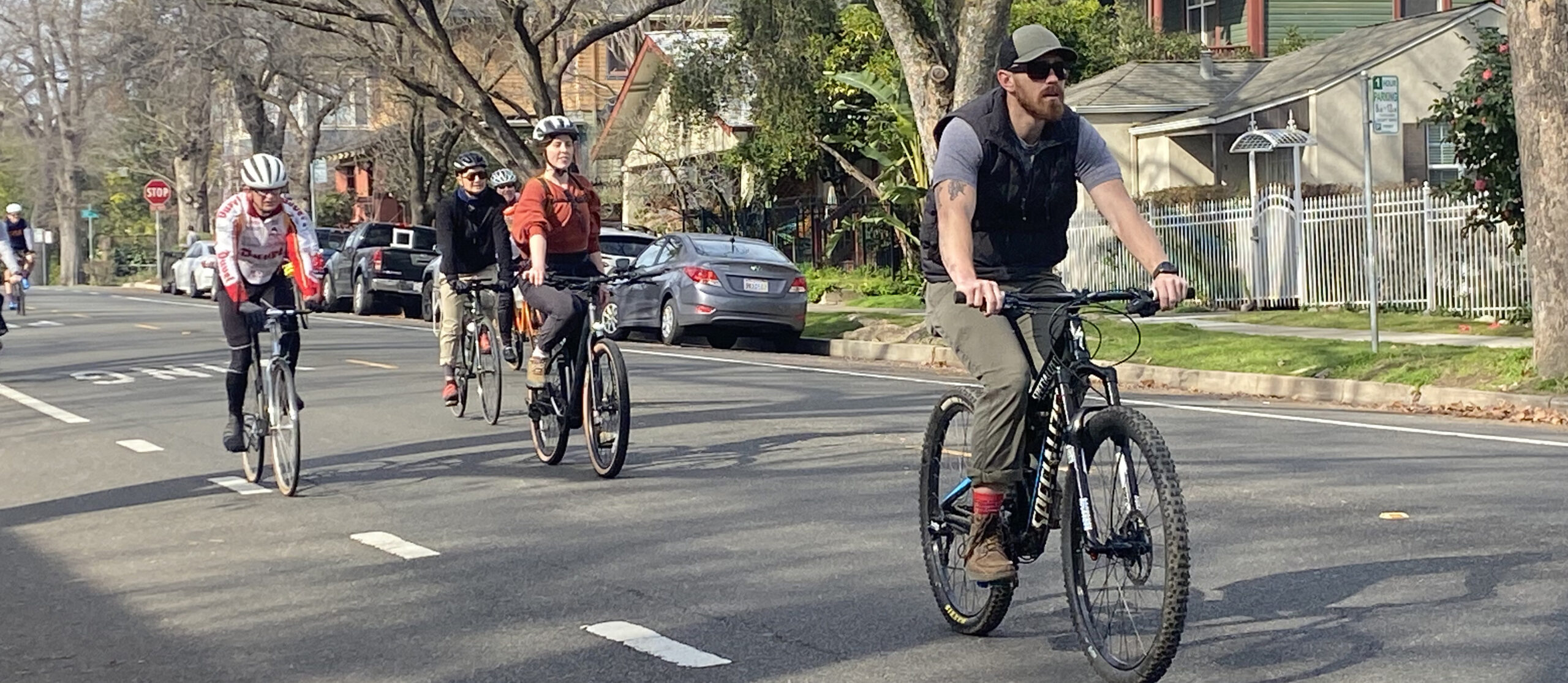 Five cyclists, 3 wearing helmets, ride down a residential street.